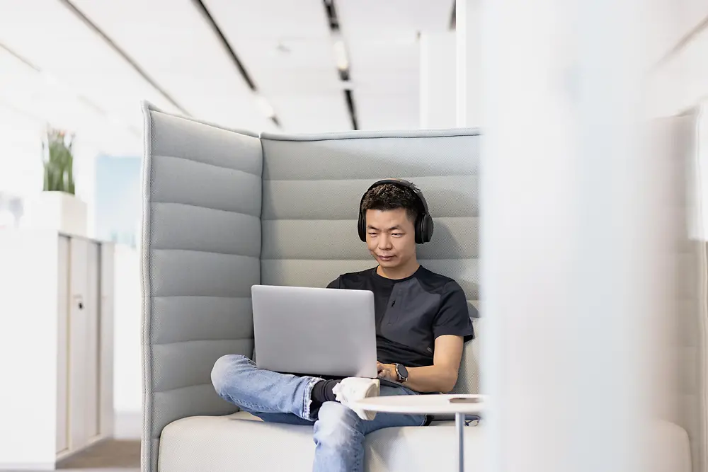 A man sits on an armchair with headphones and works on a laptop.